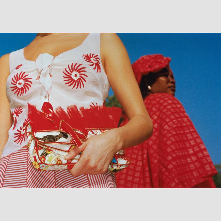A photo of two women: one woman facing camera and wearing a white and bright red dress holding a clutch; another woman in a bright red cape and hat facing away from camera.  