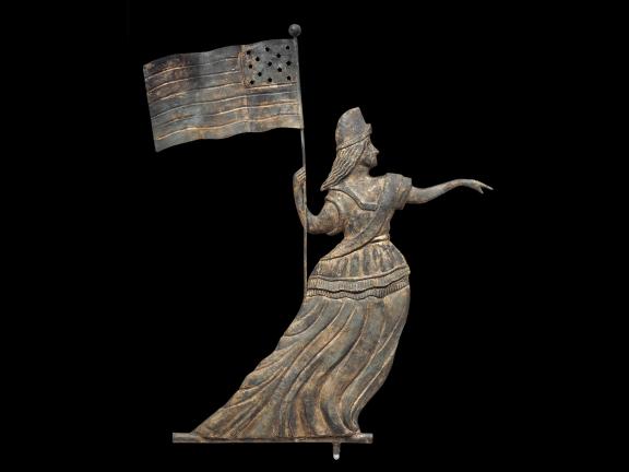 A weather vane with heavy patina that looks like a woman in a Phrygian cap and dress holding an American flag.