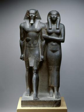 Dark grey statue of an Egyptian king and his wife with her arms wrapped around her husband.