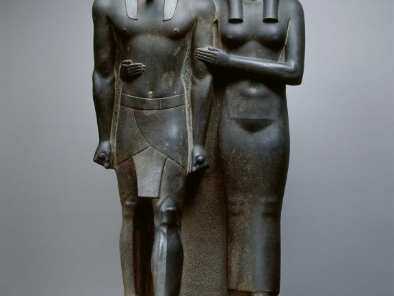 Dark grey statue of an Egyptian king and his wife with her arms wrapped around her husband.