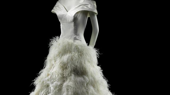 A gray mannequin wears a shiny white dress with ruffled plumes from the hip down.