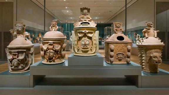 View of six Mayan urns arranged in a row inside a gallery case
