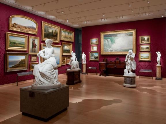 A gallery in the style of a salon display, with paintings tightly packed onto deep red walls and sculpture filling the central space