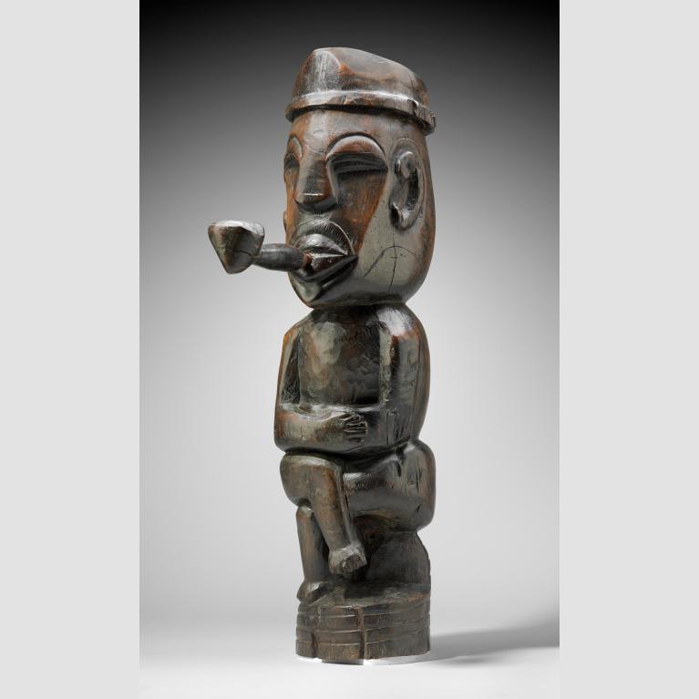 A wood sculpture of a man sitting cross-legged on a stump, wearing a hat and smoking a pipe. 