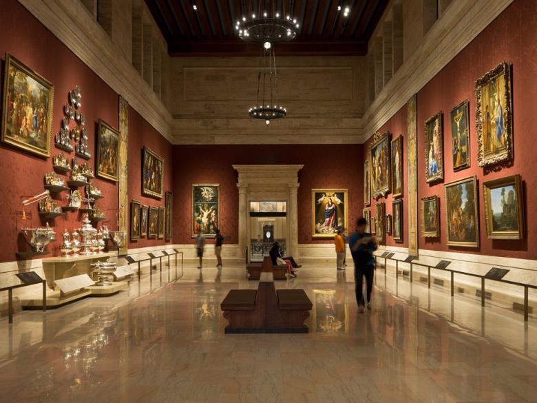 Wide view of gallery with high ceiling and reddish walled, with European paintings and silver on walls