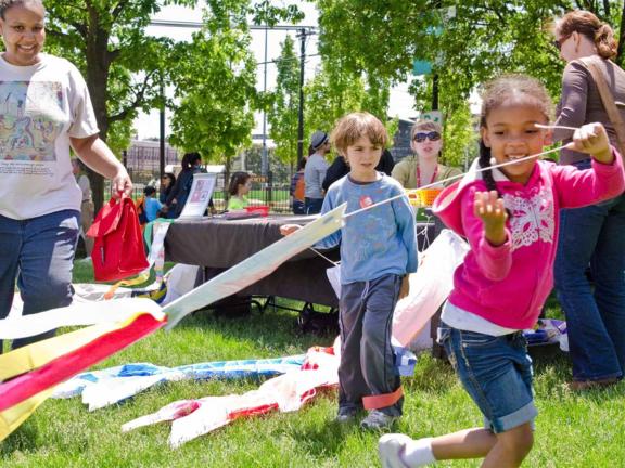 Two children play with streamers on the Huntington Lawn on a sunny day while adults and more children in the background sit at tables and participate in art-making activities