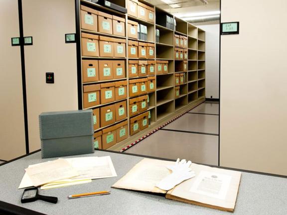 Library stacks with archival boxes, a table with old documents spread out across it.