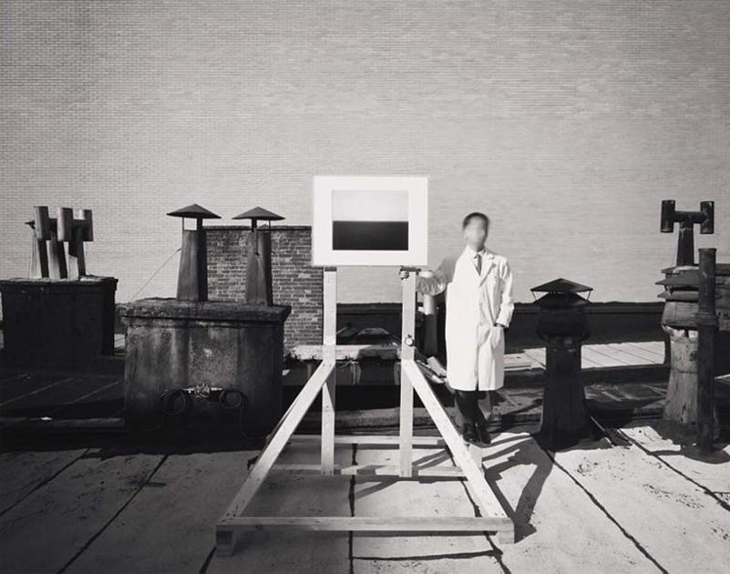A black-and-white photograph of a man in a white lab coat with a blurred face standing beside an easel on a rooftop.