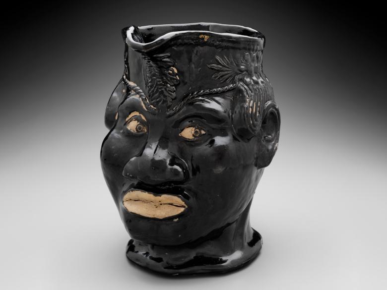 A pitcher sculpted in the form of a head, with the opening coming out of the top of a hat.  The face is a crude caricature of a black man, and the entire sculpture is painted jet black.