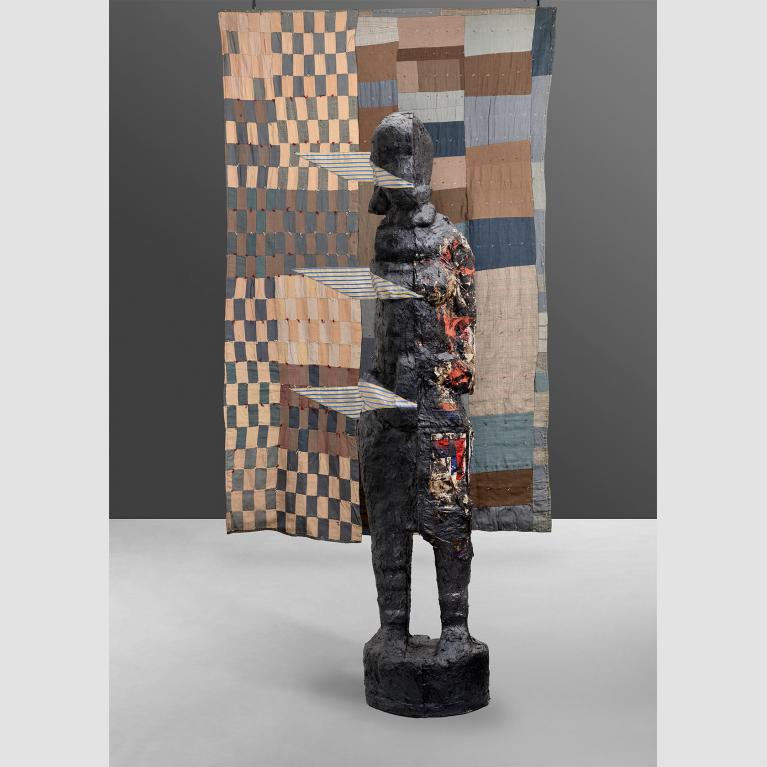 Quilt with tan, blue and brown squares and rectangles with a sculpted figure in front of it.