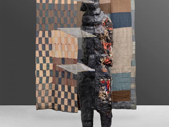 Quilt with tan, blue and brown squares and rectangles with a sculpted figure in front of it.