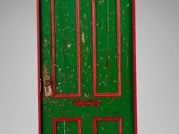 A bullet hole–riddled green door with red trim and a blue sticker reading "USA Approved."