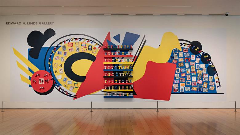 gallery installation photograph depicting colorful abstract sculpture comprised of smaller sculptures of trophies made by kids, mounted on a wall