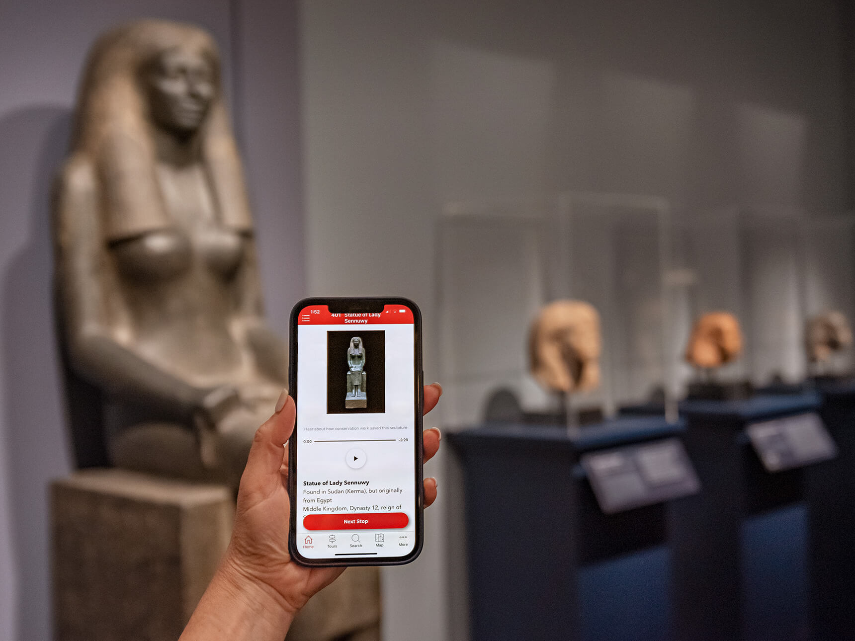 Smartphone with MFA Mobile app on screen, held up in front of Egyptian sculptures in background