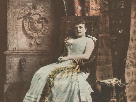 A portrait of a woman in a gown, sitting in a chair against a backdrop of curtains and blankets.
