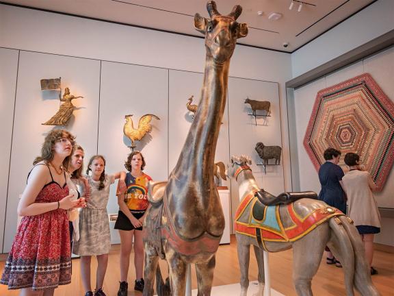 Group of young visitors looking at tall wooden sculpture of giraffe in folk art gallery