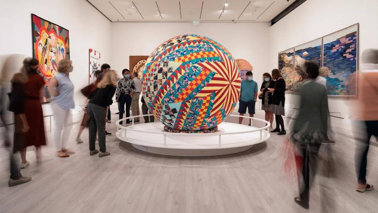 Visitors standing around large sphere made of multi-colored quilted fabrics situated on platform in center of gallery; other quilts hanging on walls of gallery