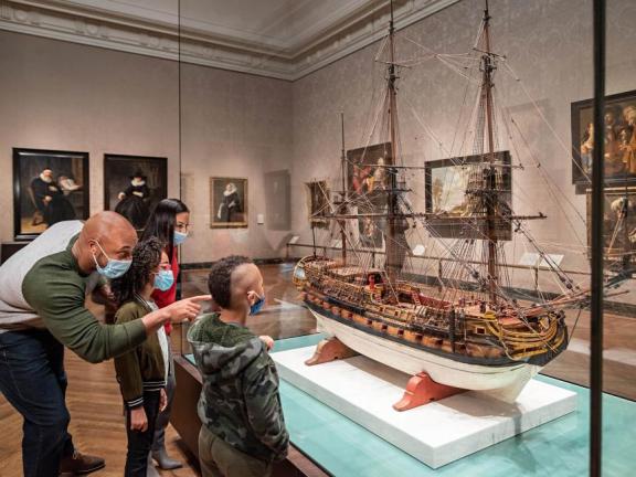 A visitor with two young children looks at a Dutch model ship behind a glass case.