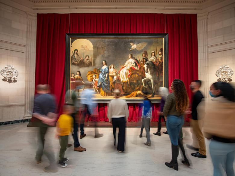 Visitors stand around a massive painting against a red curtain on a wall.