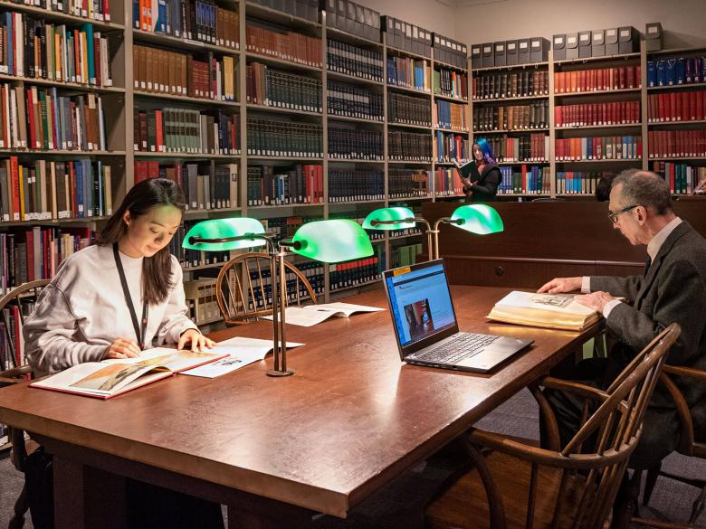Visitors reading in a library space with tall bookshelves along back wall; two seated at large table; one standing by bookshelves
