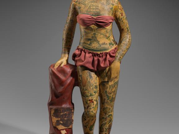 A small papier mache statuette of a heavily tattoed woman in a red bathing suit stands on a pedestal and holds a matching red garment with a crest that reads "Battleship Kate."