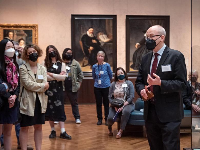 A person wearing a suit speaks to a group of people around a work of Dutch art.