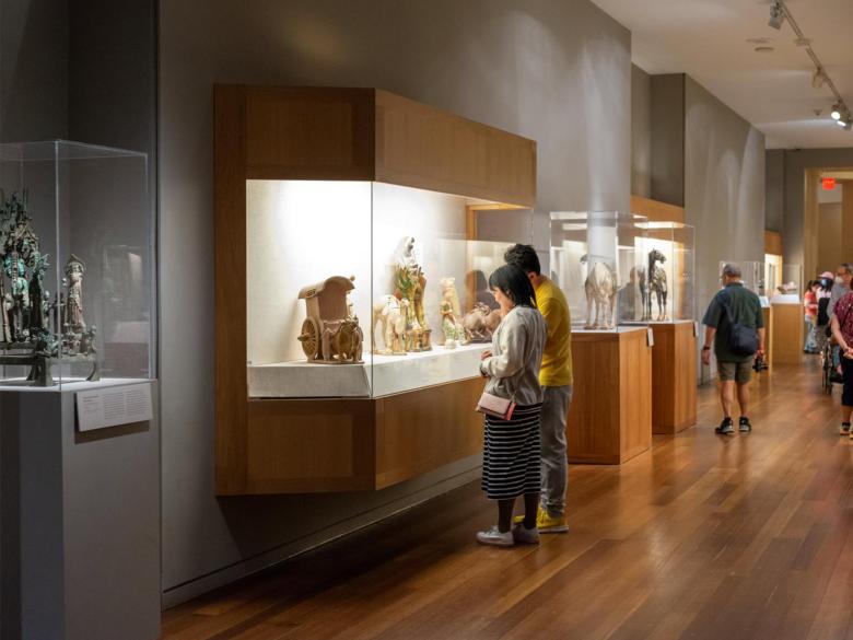 Two visitors looking at Chinese ceramic sculptures of camels and ox-drawn carts inside display case