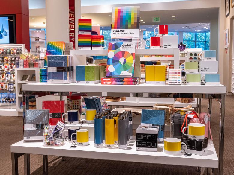 Store display featuring various Pantone-related products, including books, mugs, and notebooks, arranged on two overlapping tables