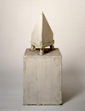 A rectangular prism sculpture topped with a pyramid-shaped sculpture on legs. 