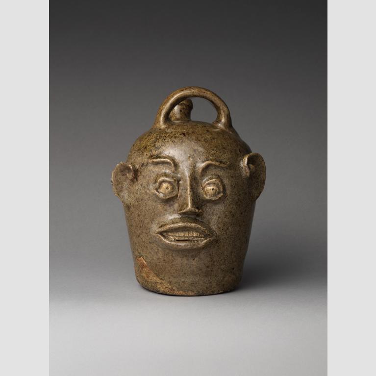 An earthenware jug with a small opening, a handle, and decorated with the features of a human face.