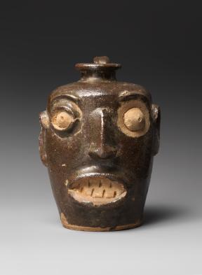 A brown and beige stoneware jug shaped into the form of a human face. 