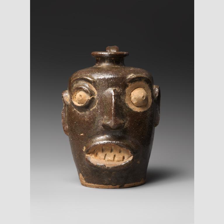 A brown and beige stoneware jug shaped into the form of a human face. 