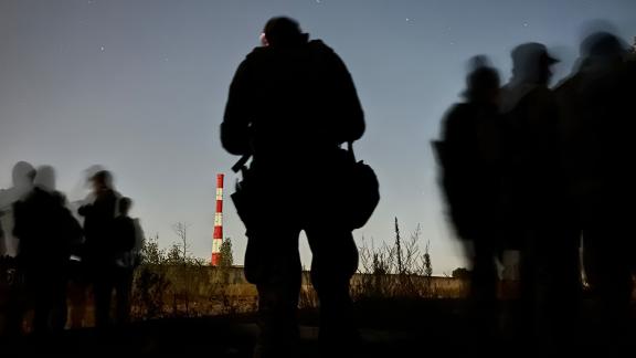 The silhouettes of Ukrainian soldiers stand out against the evening sky; a fluorescent orange and white tower sits in the distance. 
