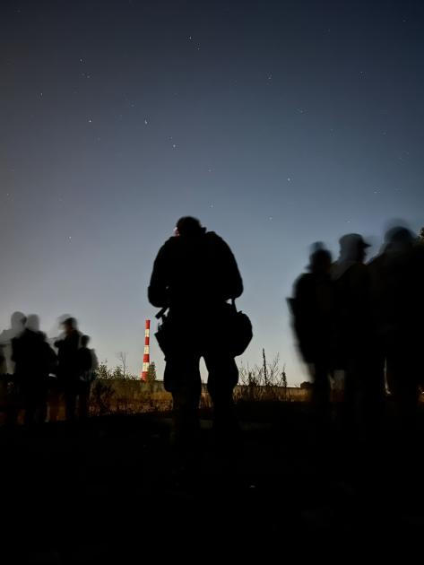 The silhouettes of Ukrainian soldiers stand out against the evening sky; a fluorescent orange and white tower sits in the distance. 