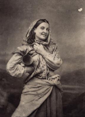 A young woman smiles for the camera with a cloth wrapped around her head and her right arm akimbo, carrying a jug. 