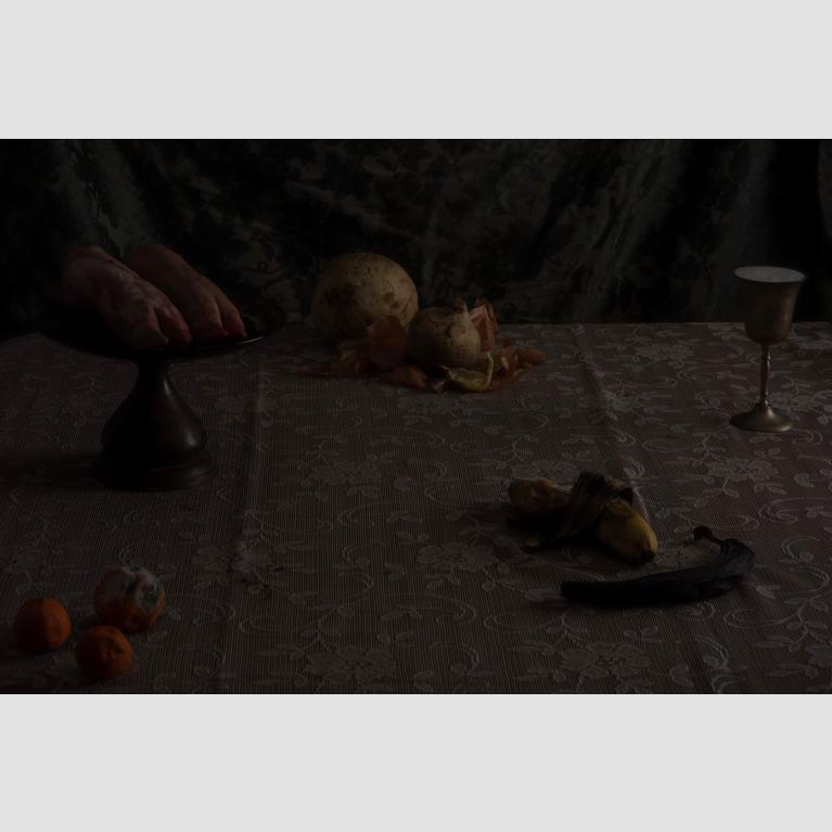 A lace-covered table set with pigs feet on a cake stand, moldy fruit and vegetables, and a goblet. 