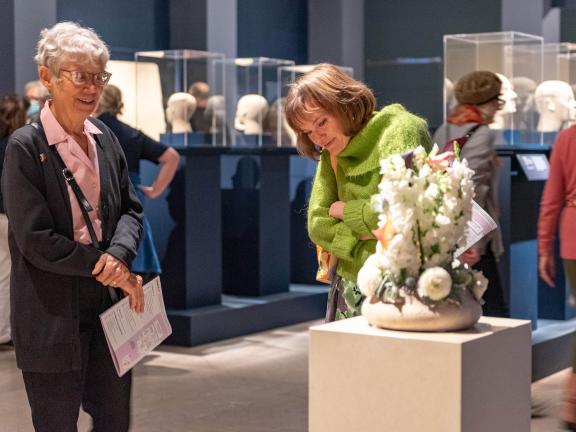 Two visitors in Egyptian gallery, looking at arrangement of white flowers in low, round vase