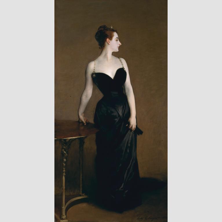 A woman in a black gown stands with her right arm leaning on a table and her left arm gathering the fabric of her dress. 