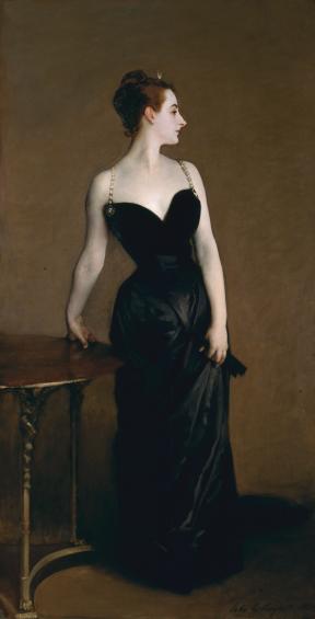A woman in a black gown stands with her right arm leaning on a table and her left arm gathering the fabric of her dress. 