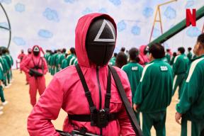 still from Squid Game depicting figure in pick jumpsuit and mask guarding over rows of people in green tracksuits