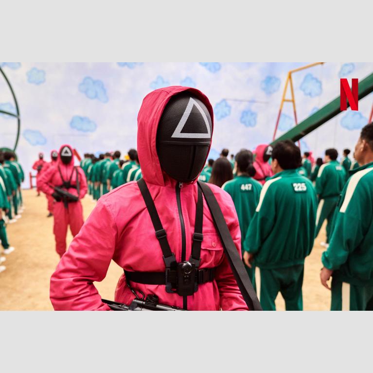 still from Squid Game depicting figure in pick jumpsuit and mask guarding over rows of people in green tracksuits