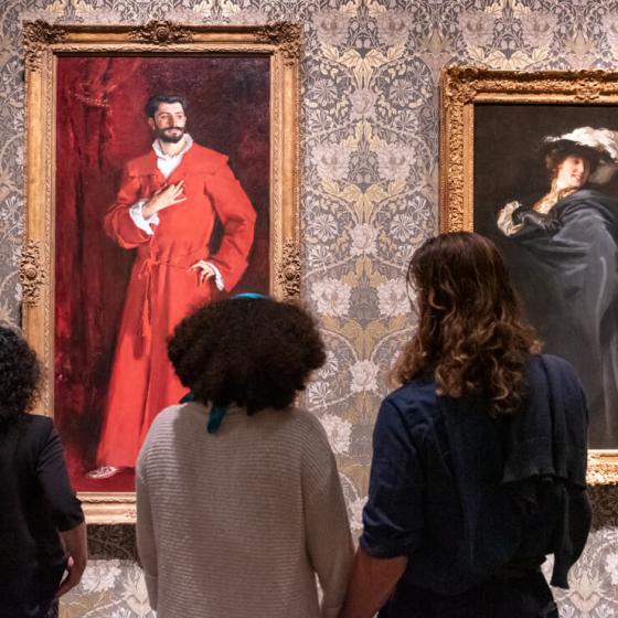 Two visitors looking at Sargent portrait paintings, of man in long red robe on left and of woman in large black cloak on right