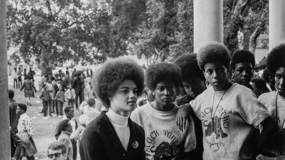 Black-and-white photo: a group of Black women look off into the distance as they stand before a crowd gathered on a lawn.