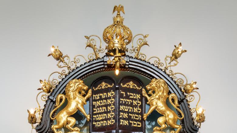 An ornate gold and wood top of a Torah ark adorned with a lights, a bird, lions, floral shapes, and two passages from the Torah. 