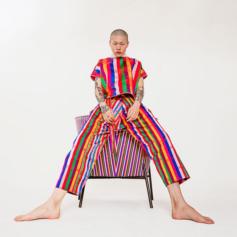 A model in a matching rainbow-striped shirt and pants leans against a piece of furniture covered in a fabric that matches his outfit.