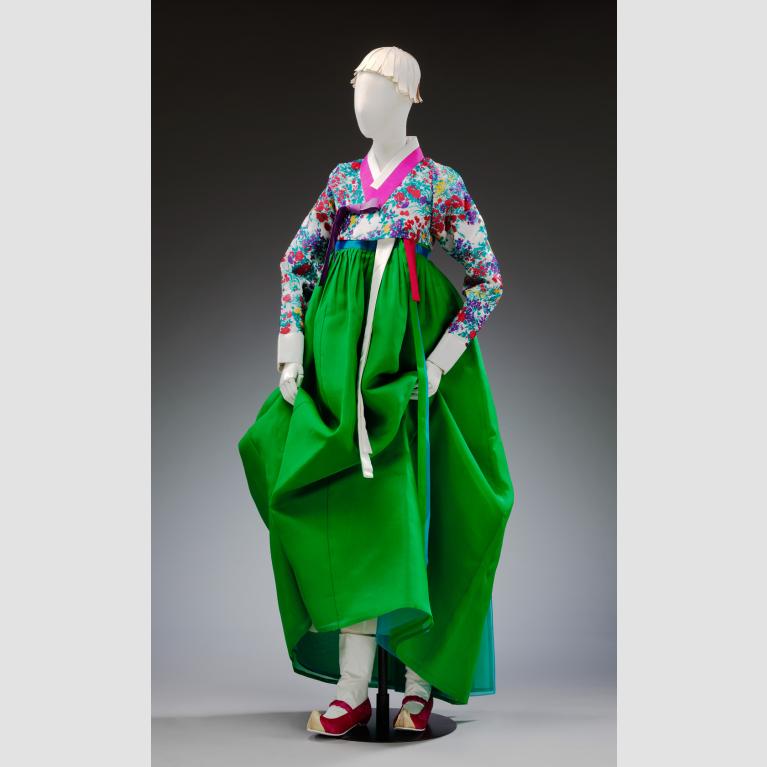 A mannequin wears a floral and green modern hanbok with red shoes and a white cloth headpiece.