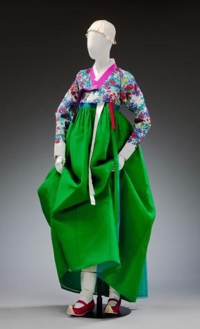 A mannequin wears a floral and green modern hanbok with red shoes and a white cloth headpiece.