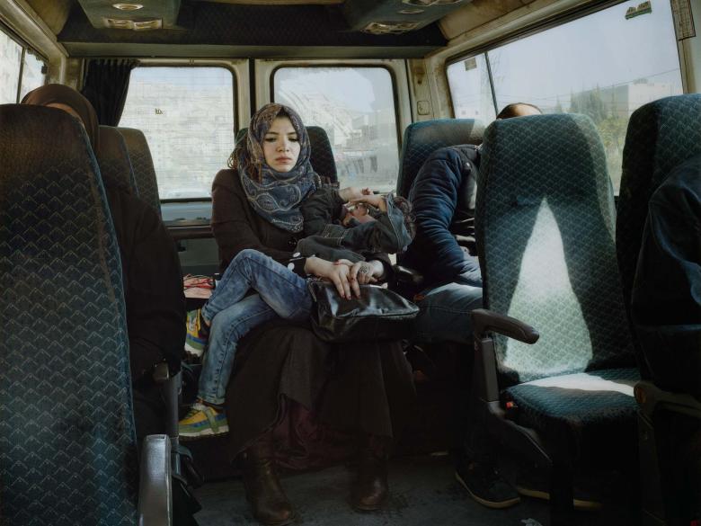 A woman with a head covering holds a writhing child in the back seat of a taxi cab as sunlight streams in through the window.