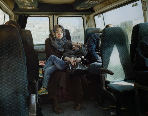A woman with a head covering holds a writhing child in the back seat of a taxi cab as sunlight streams in through the window.