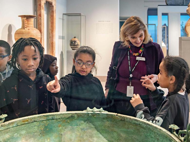 Group of kids, with one pointing, with MFA instructor looking at ancient Greek bronze urn in gallery case